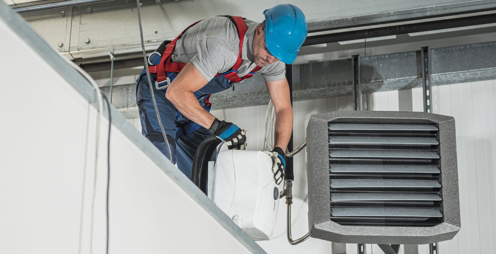 Heating engineer in a hard hat and harness installing a warm air unit heater at height in a warehouse